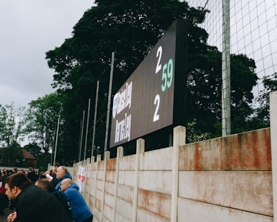 A large digital scoreboard at an outdoor sports venue displays a score of 2-2. Spectators are gathered, observing the event with interest. The scene is enclosed by a tall metal fence and surrounded by lush green trees, indicating a natural setting. Overcast skies add a muted tone to the environment.