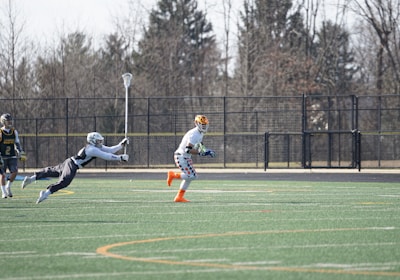 A group of athletes engaged in a sports game on a green field. One player is running forward with possession of the ball, wearing colorful socks and a checkered uniform, while another player in dark clothing dives to intercept or tackle. Other players are present in the background. Leafless trees and a metal fence surround the field.