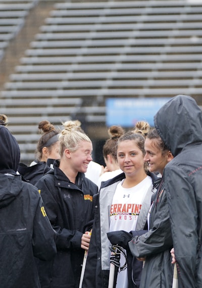A group of people, likely athletes, are gathered together on a sports field. They are wearing black jackets with some individuals dressed in white sports uniforms underneath. A few people have their hair tied up in buns, and most are engaged in conversation. The background shows tiered stadium seats, suggesting the setting is an outdoor sports venue.