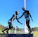 A sculpture depicting two athletes in mid-action is prominently displayed against a clear blue sky. One figure is reaching out to help the other, illustrating a moment of sportsmanship. The bronze statues stand on a black pedestal with an inscription, situated in an open green field with trees and sports facilities in the background. A tall floodlight tower looms overhead, adding to the outdoor sports setting.