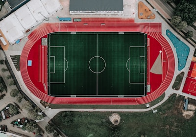 An aerial view of a large sports complex featuring a central green football field surrounded by a red running track. Adjacent to the track, there is a parking area, buildings, and pathways. Nearby, there are additional sports courts in different colors, including a blue swimming pool and orange basketball courts.