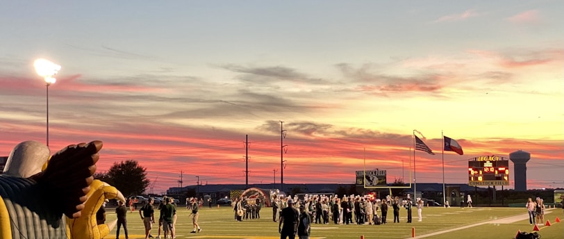 A sports field with a football game unfolding under a vibrant sunset sky, featuring a large inflatable mascot and a scoreboard displaying the score. Players and coaches are visible on the green field, surrounded by a track and some flags waving in the breeze.