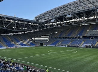 A modern sports stadium with a large, green soccer field and surrounding seating areas. Blue and yellow seats are visible, with some spectators scattered across the stands. The stadium is partially covered with a steel lattice roof, showing a clear blue sky. Several security personnel in high-visibility vests are present.