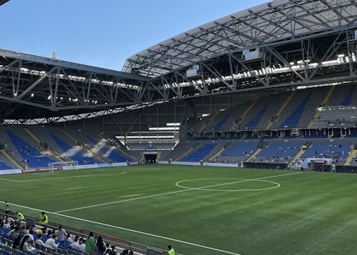A modern sports stadium with a large, green soccer field and surrounding seating areas. Blue and yellow seats are visible, with some spectators scattered across the stands. The stadium is partially covered with a steel lattice roof, showing a clear blue sky. Several security personnel in high-visibility vests are present.